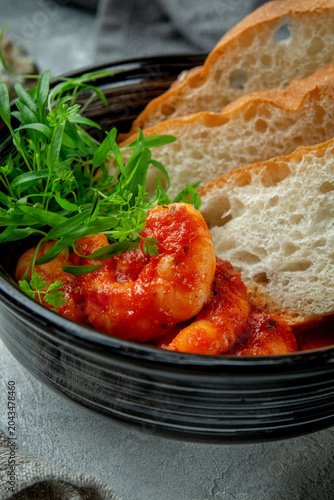Close-up of spicy shrimp in tomato sauce with crispy bread and fresh herbs in a black bowl on rustic table