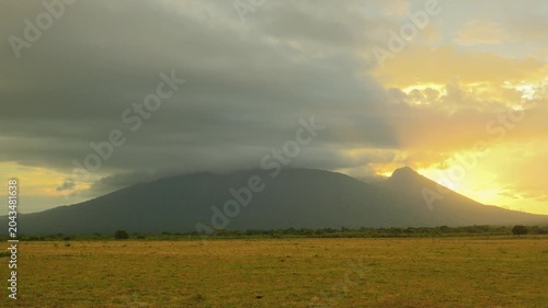 Dramatic timelapse of Mount Baluran volcano in Situbondo, Indonesia under fast moving storm clouds at golden sunset with vast green savanna field.