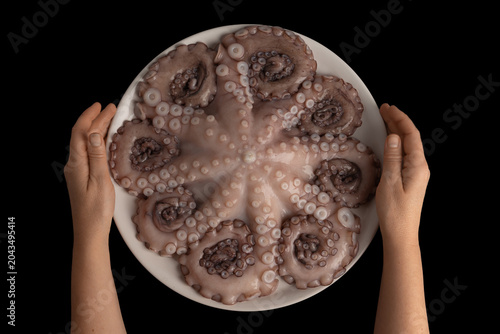 Person's hands presenting a whole raw octopus with visible tentacles and suckers on a white plate, ready for preparing a healthy gourmet seafood meal against a black background