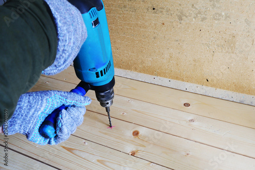 Close-up shot of a woodworking process. A person, wearing grey knitted work gloves, is using a blue and black cordless electric drill to create a pilot hole in a light-colored pine wooden floorboard