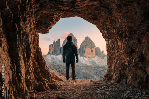 Bild auf Leinwand Male tourist admiring the Tre Cime Peak from rocky cave during evening in Dolomi