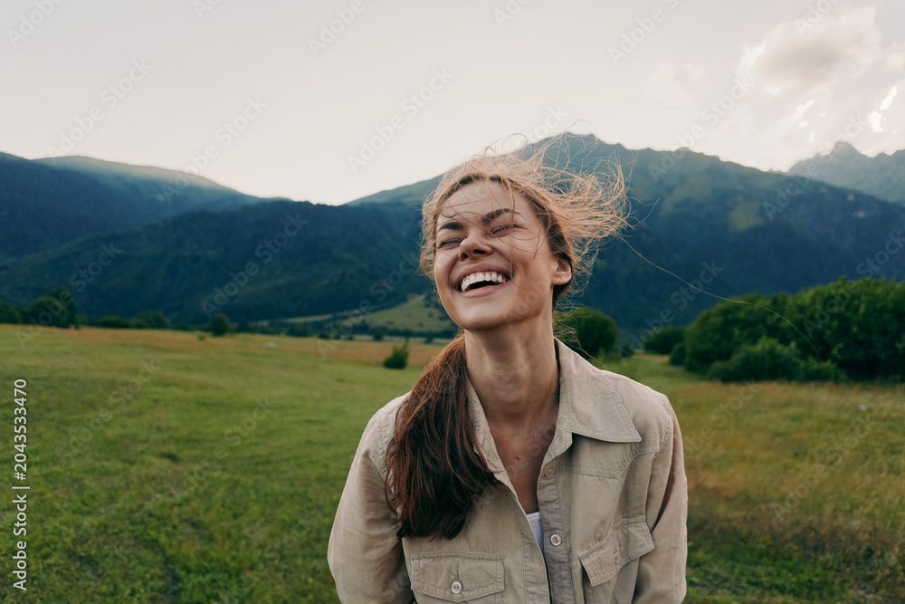Fototapeta premium Cheerful woman outdoors, smiling with eyes closed, sunlit moment in a field near mountains, casual jacket, light breeze, natural colors, lush greenery, carefree mood today