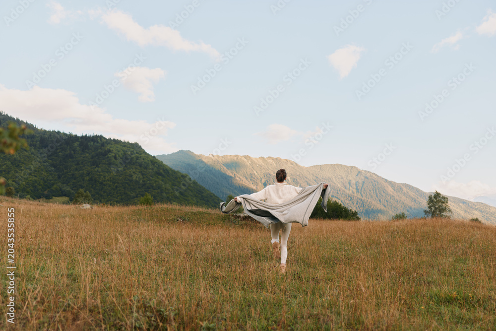 Fototapeta premium Young woman stands in a sunlit field with arms outstretched, a flowing shawl catching the breeze, while distant mountains frame a peaceful, expansive countryside scene.