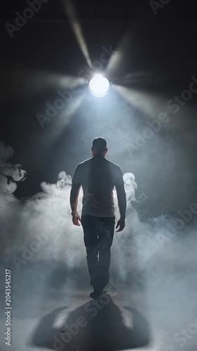 Man walking away into bright spotlight with atmospheric smoke on dark background, tracking shot