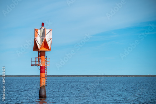 Lighthouse. Orange maritime navigation light beacon on a steel pole in the blue sea water against a clear sky, minimalist coastal shipping lane safety sign with copy space.