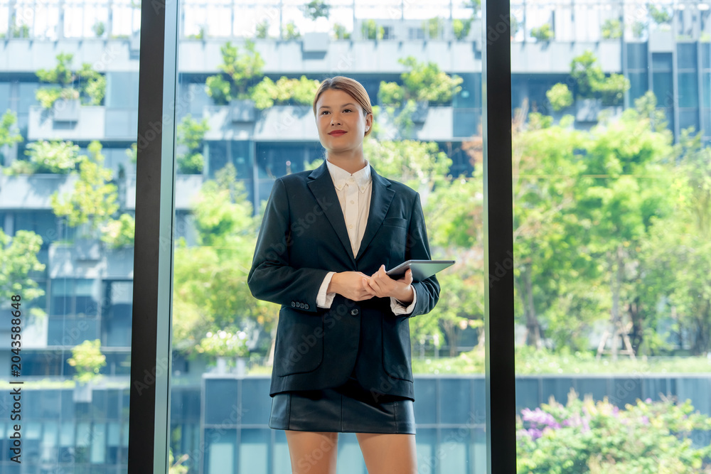 Fototapeta premium Portrait of a confident and successful young businesswoman in black suit holding a digital tablet near modern office window with visionary expression