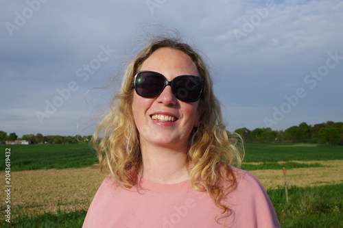 Woman with long blonde hair and sunglasses, smiling in an outdoor setting. She is wearing a pink top and appears happy. 