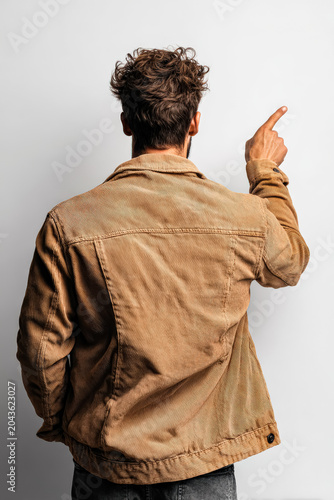 A man dressed in neutral clothing is seen from the back, pointing with his finger. The image captures the subject in a studio setting with a plain white background, emphasizing the action of pointing.