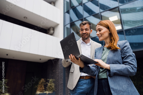 Photos Two diverse business professionals standing outside a modern glass office building, looking at a laptop screen and discussing a project during a meeting