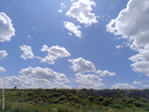 Scenic view of a vast, brilliant blue sky filled with numerous white, fluffy cumulus clouds, stretching above a natural grassy hillside on a bright, sunny day. The sun is shining brightly