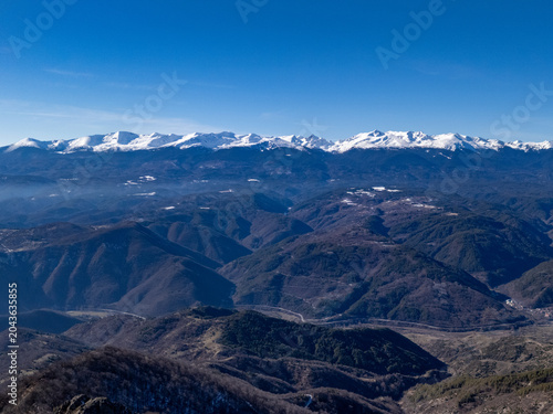 A vast, rugged mountain landscape unfolds with layers of dark, forested hills leading to distant, snow-covered summits under a brilliant azure sky