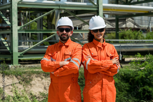 Multiracial male technician with female engineer pose arms crossed at refinery pipeline site, representing petrochemical safety culture, compliance readiness, field workforce confidence portrait