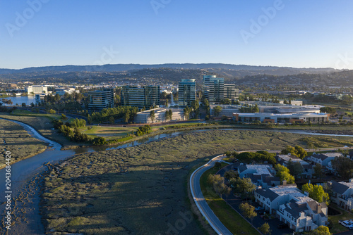 Redwood City shoreline with corporate office campuses and suburban homes beside marsh wetlands and estuary waters under a clear sunny sky, merging urban development and nature