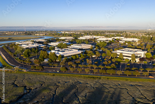 Redwood LIFE Center, an innovative Silicon Valley tech campus featuring modern office buildings and parking lots, borders serene wetlands and the Redwood Shores lagoon under clear skies