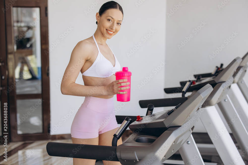 Naklejka premium Sporty young woman in gym wearing sportswear, standing on treadmill with shaker, practicing fitness for health