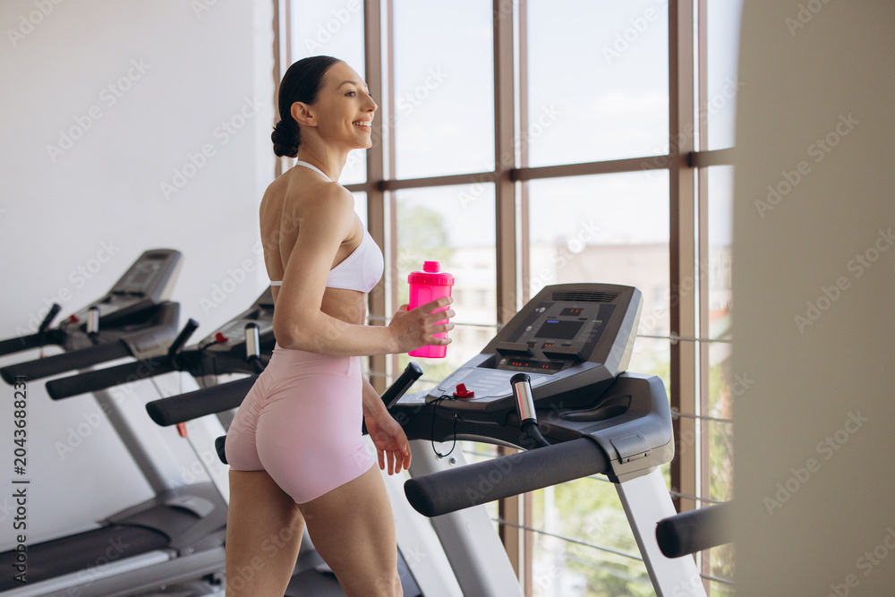 Fototapeta premium Athletic woman exercising on a treadmill at the gym, staying active and hydrated with a pink water bottle