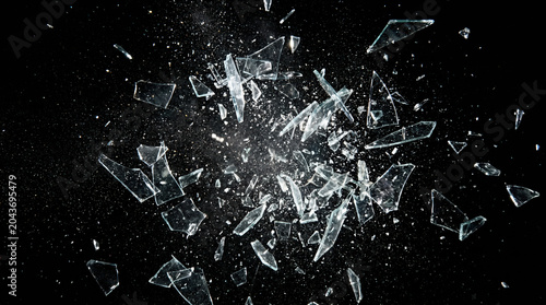 Explosion of shattered glass shards and tiny fragments flying through the air against a solid black background, captured in a high-speed motion freeze.