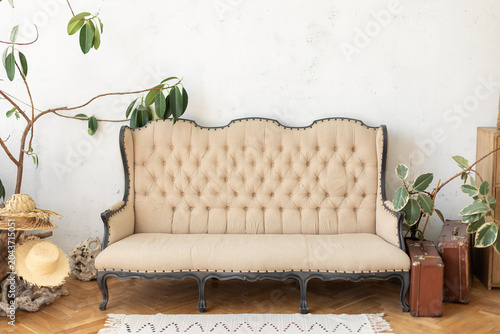 Elegant vintage tufted sofa in cozy beige fabric against white concrete wall. Modern bohemian style living room interior decorated with potted ficus plants, straw hats and retro brown suitcases.