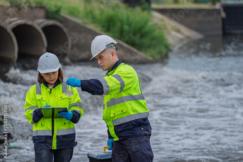 Fotografie Environmental engineers collecting water samples for laboratory analysis and quality control monitoring near industrial drainage pipes to ensure safety and ecological sustainability standards