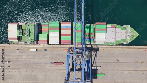 Top-down aerial perspective showing a large gantry crane moving along the quay while a massive container ship is docked in the harbor, with trucks moving cargo on the industrial pier