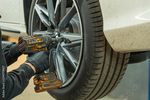 Gloved hands use a cordless impact wrench to tighten wheel bolts on a raised car wheel in a workshop, with a close view of the tire, rim, and vehicle body