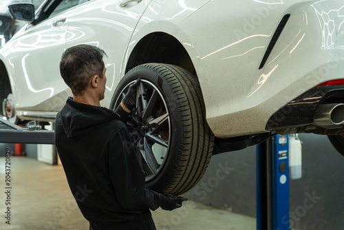 Technician working beside a white car on a hydraulic lift while checking the rear wheel in an automotive repair garage with bright indoor lighting