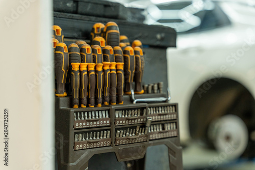Assorted screwdrivers and precision bits stored in a neat black organizer inside a garage workshop, with a blurred vehicle wheel in the background and a