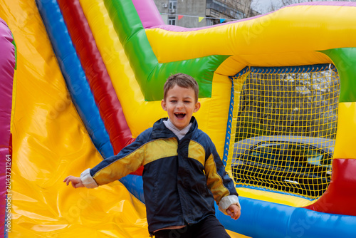 A joyful boy bursts out of a colorful bouncy castle with a huge laugh, arms spread wide, wearing a blue and yellow jacket at an outdoor playground or festival event