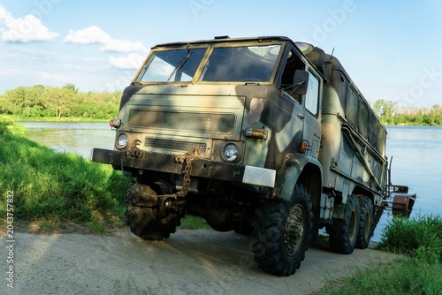 A horizontal photo of an old military truck parked on the bank of a river.