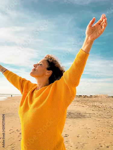 Obraz na plátně Woman in a yellow sweater standing on a sunny beach with arms raised, eyes close
