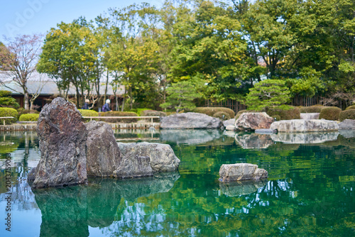 Tableau sur toile A serene Japanese garden featuring a tranquil emerald-green pond with large weathered rocks and stones arranged in the water