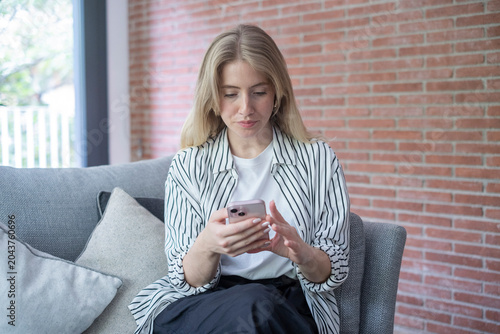 Obraz na płótnie Young woman sitting on a sofa in a modern living room, focused on her smartphone as she scrolls and types, relaxed casual style with striped shirt and neutral home decor background