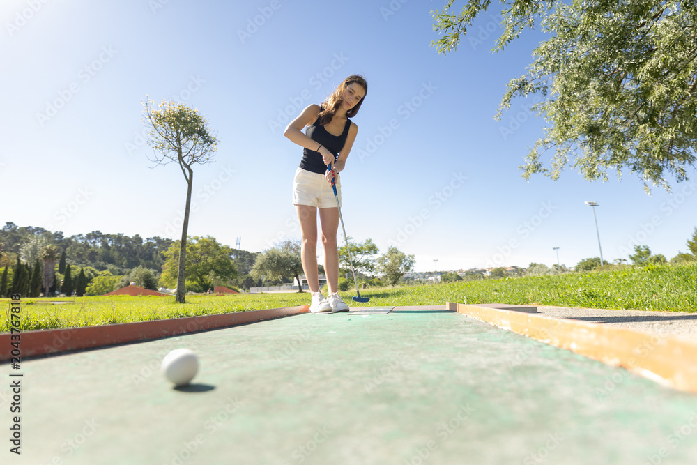 Naklejka premium Woman plays mini golf on a sunny day in a park with green grass and trees around the course in the afternoon