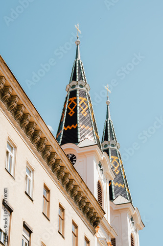 Upward view of two ornate church towers featuring steep spires topped with crosses and decorative patterned tile roofs. The pale stone facade of the adjacent