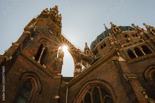 A dramatic low-angle view of an ornate brick cathedral showing tall clock tower and domed roof details against a pale sky. Sunlight creates a starburst flare