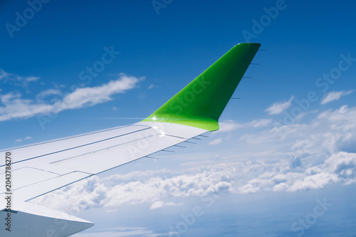 View from a passenger window showing an aircraft wing and bright green winglet above a sea of white clouds under a vivid blue sky. Smooth metallic wing