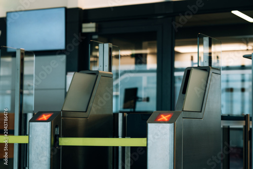 A row of stainless steel automated turnstile gates with glass panels at a transit entrance. Two gate terminals in the foreground display illuminated red X