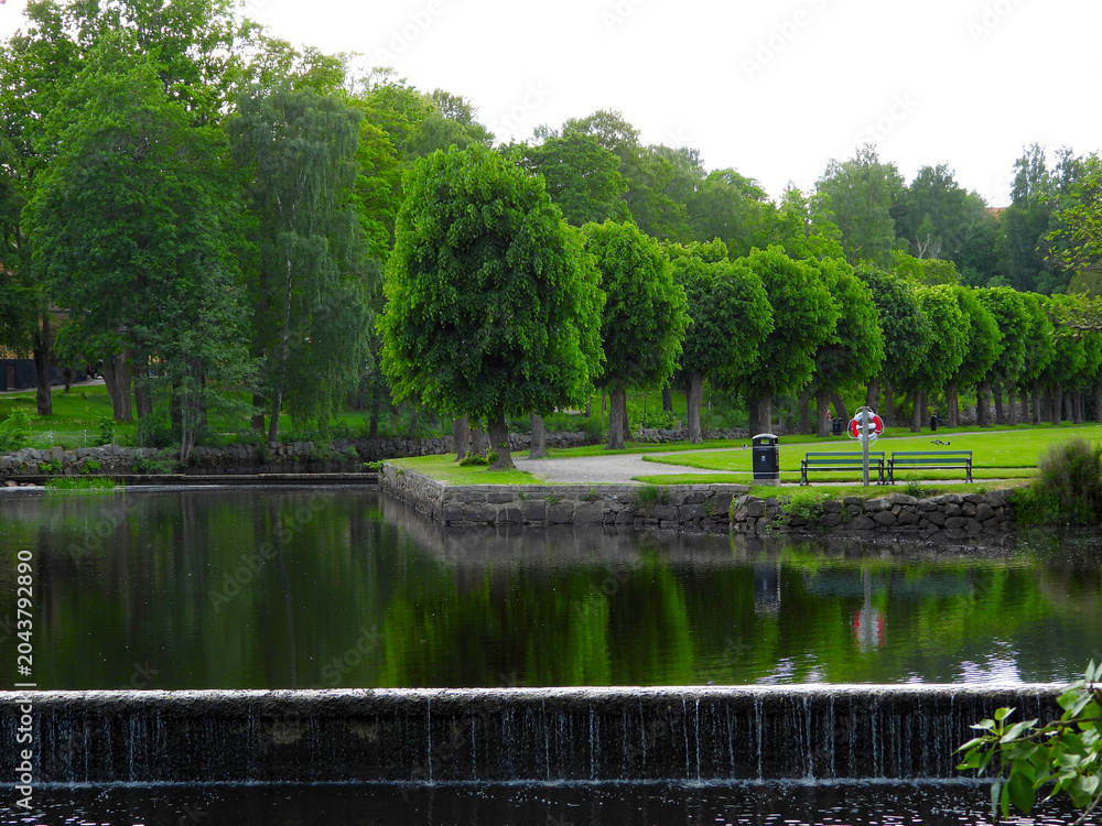 Fototapeta premium A beautiful scenic landscape in a city park in Finspång, Sweden, featuring a calm lake surrounded by green grass and natural vegetation. The peaceful environment reflects a typical Scandinavian park a