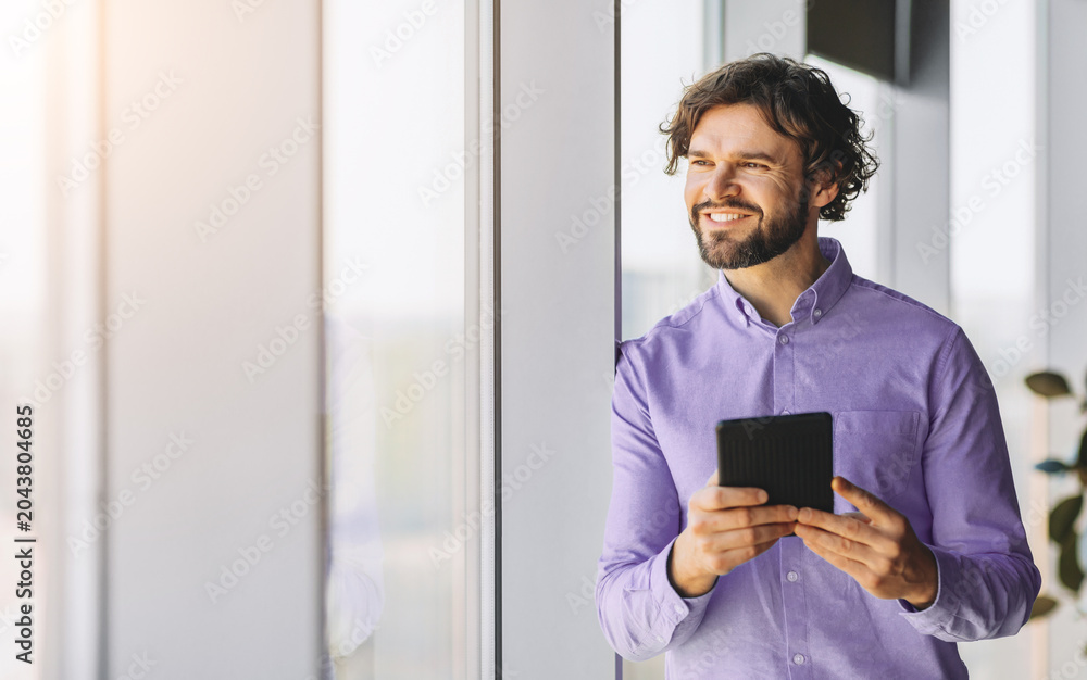 Fototapeta premium A man stands by a large window in a bright office space. He is smiling and looking outside while holding a tablet. Sunlight shines through the window creating a warm atmosphere