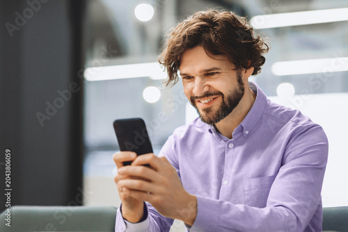 Fotografía A man sits comfortably in a modern indoor area, smiling at his smartphone