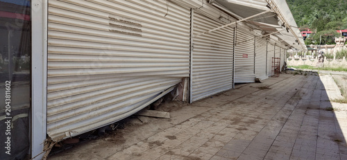 Quadro su tela Flood-damaged abandoned shops with collapsed metal shutters and debris in outdoo