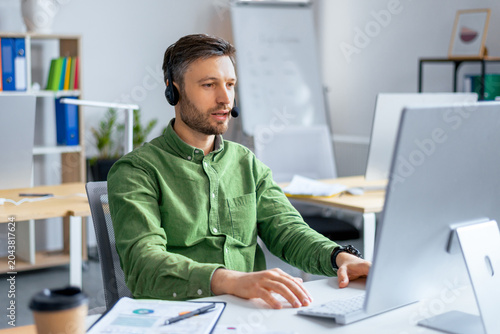Tableau sur toile Focused Caucasian Man Wearing Headset Working On Computer, Engaged In Task At Modern Office Environment