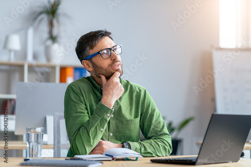 Quadro su tela Focused Caucasian Man In Green Shirt Thinking While Working On Laptop At Modern Office, Sitting At Desk With Notebook And Glass Of Water