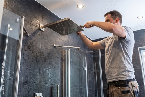 A man is installing a modern square shower head in a contemporary bathroom with dark tiled walls.