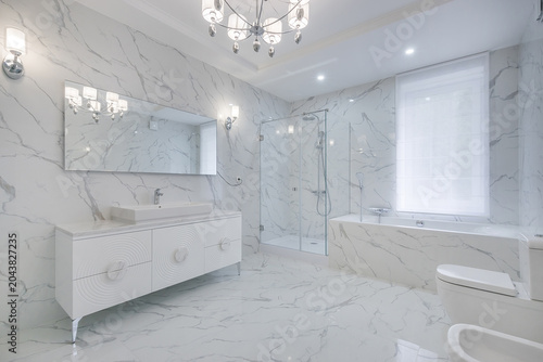 The magnificent design of the bathroom, decorated with light marble tiles. Shower cabin and bathtub by the window.