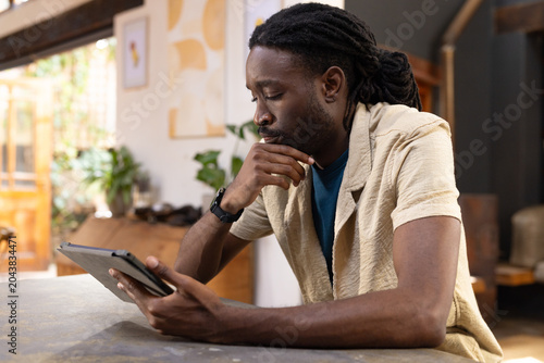 African American man sitting at table at home holding tablet, wearing beige shirt and smartwatch