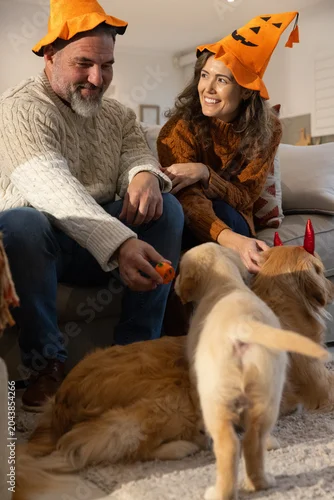 Fototapeta Couple sitting on sofa wearing pumpkin hats, holding pumpkin toy, petting dog with red horns
