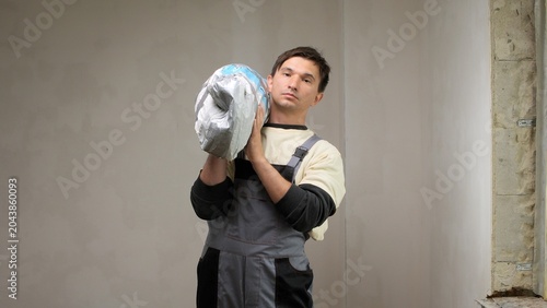 Bild auf Leinwand Construction worker in overalls carrying a heavy bag of cement or putty on his s