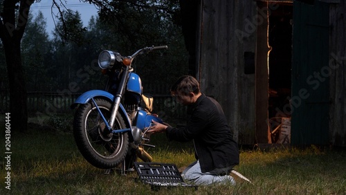 Obraz na plátně Young man kneeling in a grassy field, adjusting an old blue motorcycle engine du