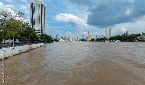 Fotografie A wide view of the Chao Phraya River in Bangkok, Thailand, with a modern cityscape and skyscrapers in the background under a cloudy sky